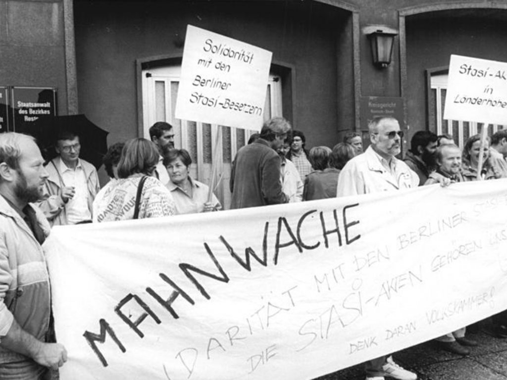 Eine Menschenmenge protestiert vor einem Gebäude der Staatsanwaltschaft Rostock. Die Menschen halten ein großes Banner mit der Aufschrift: Mahnwache. Solidarität mit den Berliner Stasi-Besetzern. Die Stasi-Akten gehören uns. Denk daran Volkskammer. Im Hintergrund werden zwei Plakate gehalten mit den Beschriftungen: Solidarität mit den Berliner Stasi-Besetzern. Und: Stasi-Akten in Länderhoheit.