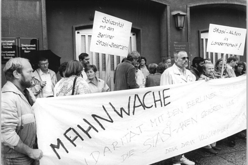 Eine Menschenmenge protestiert vor einem Geb&auml;ude der Staatsanwaltschaft Rostock. Die Menschen halten ein gro&szlig;es Banner mit der Aufschrift: Mahnwache. Solidarit&auml;t mit den Berliner Stasi-Besetzern. Die Stasi-Akten geh&ouml;ren uns. Denk daran Volkskammer. Im Hintergrund werden zwei Plakate gehalten mit den Beschriftungen: Solidarit&auml;t mit den Berliner Stasi-Besetzern. Und: Stasi-Akten in L&auml;nderhoheit.
