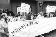 Eine Menschenmenge protestiert vor einem Gebäude der Staatsanwaltschaft Rostock. Die Menschen halten ein großes Banner mit der Aufschrift: Mahnwache. Solidarität mit den Berliner Stasi-Besetzern. Die Stasi-Akten gehören uns. Denk daran Volkskammer. Im Hintergrund werden zwei Plakate gehalten mit den Beschriftungen: Solidarität mit den Berliner Stasi-Besetzern. Und: Stasi-Akten in Länderhoheit.
