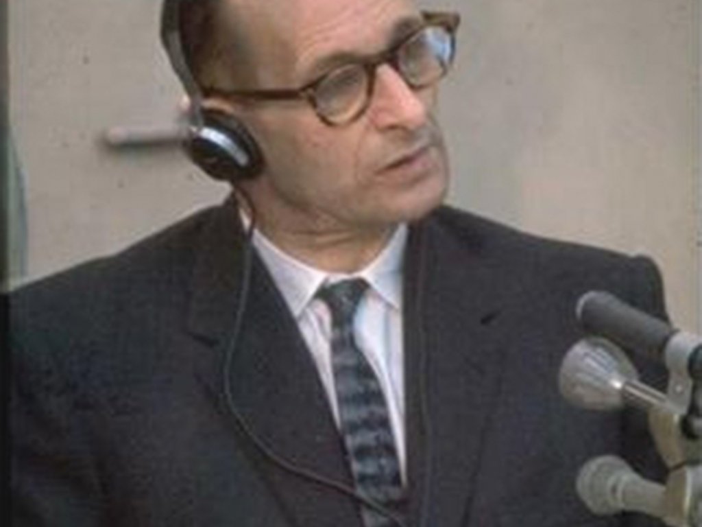 Adolf Eichmann trägt Kopfhörer und hat den Kopf leicht schräg gelegt, vor ihm stehen Mikrofone.