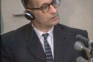 Adolf Eichmann trägt Kopfhörer und hat den Kopf leicht schräg gelegt, vor ihm stehen Mikrofone.