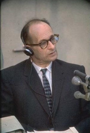 Adolf Eichmann trägt Kopfhörer und hat den Kopf leicht schräg gelegt, vor ihm stehen Mikrofone.