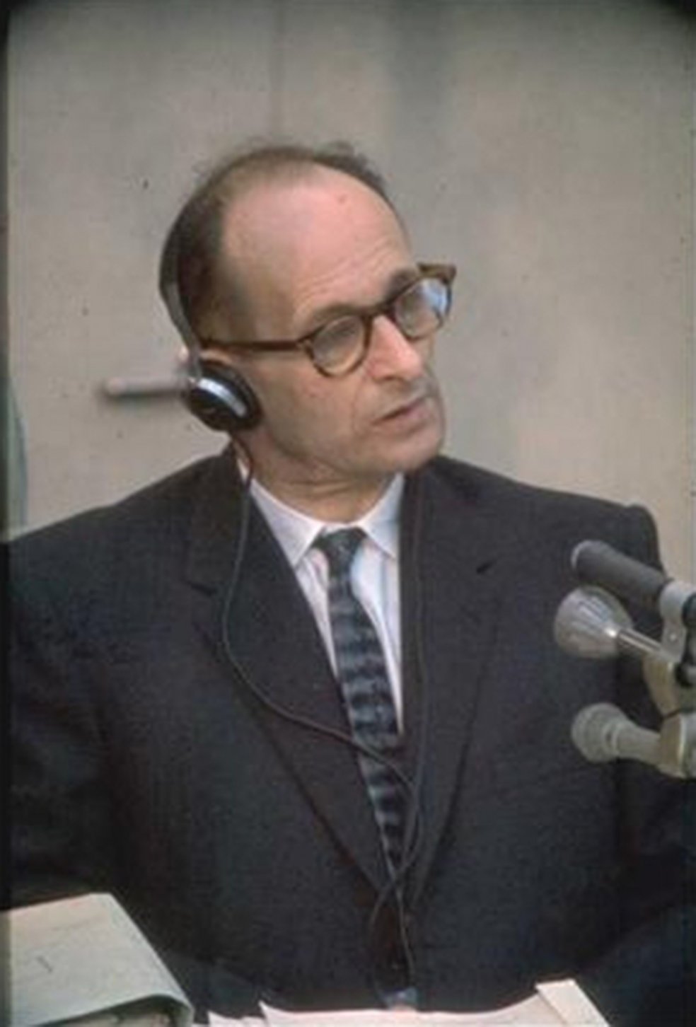 Adolf Eichmann tr&auml;gt Kopfh&ouml;rer und hat den Kopf leicht schr&auml;g gelegt, vor ihm stehen Mikrofone.