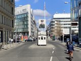 Checkpoint Charlie an einem sonnigen Tag, rechts Sektorenschild mit Aufschrift: You are leaving the american sector, darunter auf russisch und französisch. Mittig Kontrollbaracke mit Sandsäcken auf einer Verkehrsinsel. In einigen Metern Höhe zeigt ein Leuchtkasten ein überlebensgroßes Foto eines russischen Soldaten.