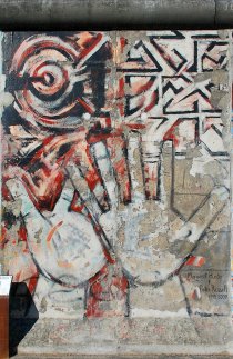 The lower parts of the weathered painting on conrete shows the outline of two overlapping hands with outstreched fingers reaching upwards. In the upper portion of the painting, one sees geometric patterns.