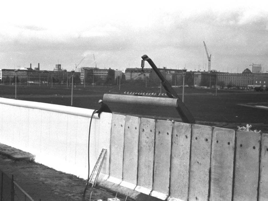 Ein Angehöriger des Bautrupps befestigt eine Rohrauflage auf der Grenzmauer. Daneben befindet sich ein Kran. Vor der Baustelle steht ein mobiler Sicherungszaun und ein Grenzsoldat. Im Hintergrund ist der Grenzstreifen mit Fahrzeugsperren sichtbar.
