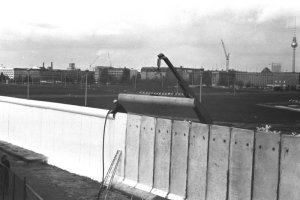 Ein Angeh&ouml;riger des Bautrupps befestigt eine Rohrauflage auf der Grenzmauer. Daneben befindet sich ein Kran. Vor der Baustelle steht ein mobiler Sicherungszaun und ein Grenzsoldat. Im Hintergrund ist der Grenzstreifen mit Fahrzeugsperren sichtbar.
