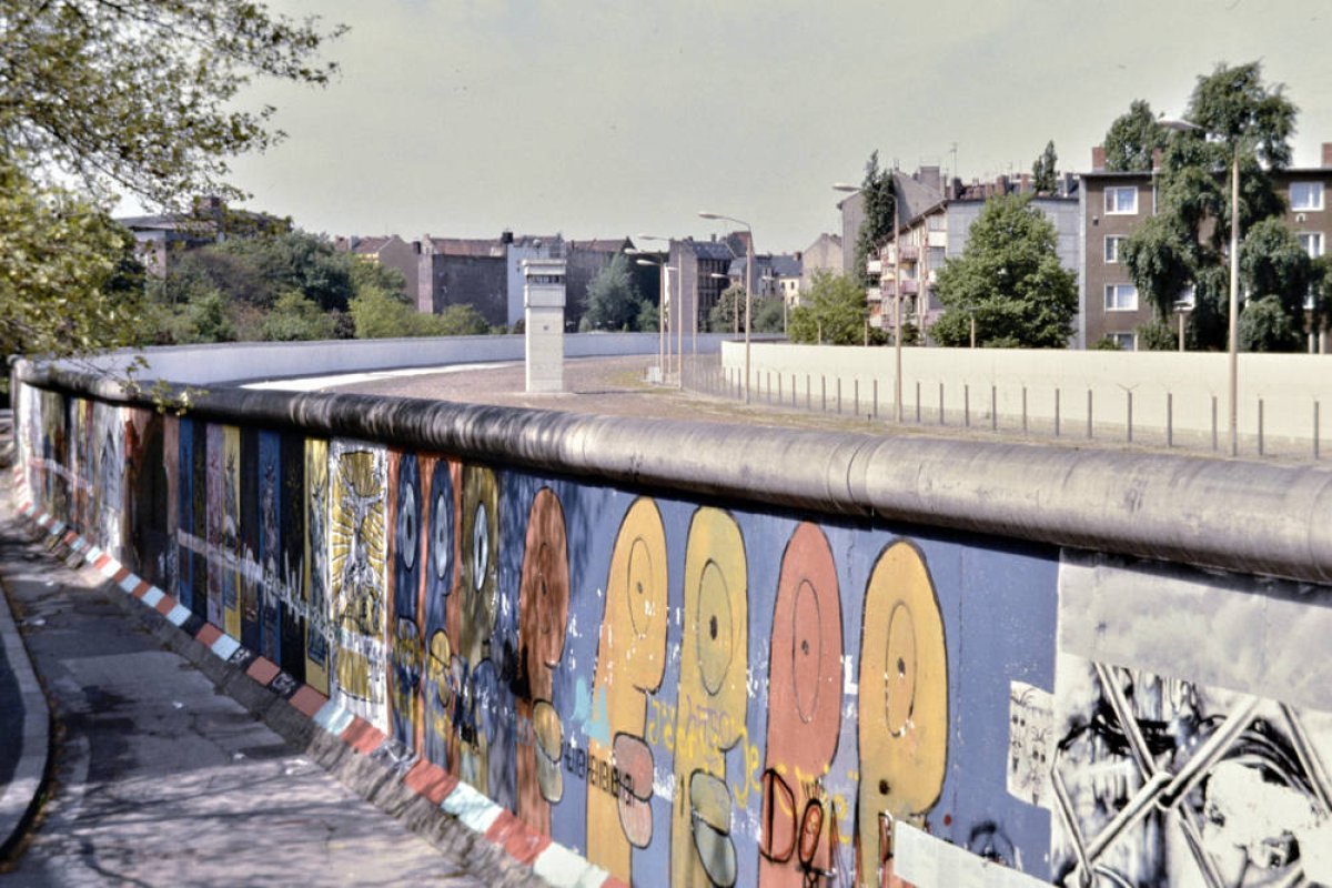 Der untere Bildteil zeigt Thierry Noirs bunte Köpfe auf der Mauer. Oben sieht man in den Grenzstreifen, im Hintergrund steht ein Wachturm. Der untere Bildteil zeigt Thierry Noirs bunte Köpfe auf der Mauer. Oben sieht man in den Grenzstreifen, im Hintergrund steht ein Wachturm.