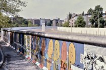 Der untere Bildteil zeigt Thierry Noirs bunte Köpfe auf der Mauer. Oben sieht man in den Grenzstreifen, im Hintergrund steht ein Wachturm.