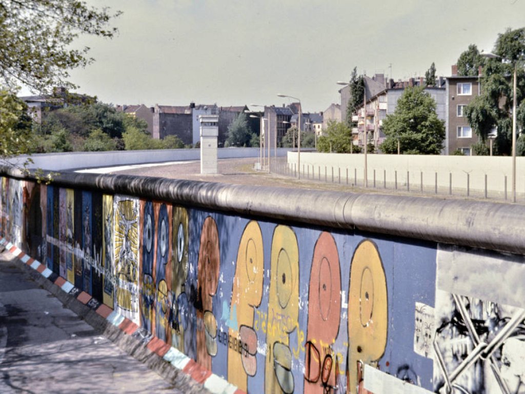 Der untere Bildteil zeigt Thierry Noirs bunte Köpfe auf der Mauer. Oben sieht man in den Grenzstreifen, im Hintergrund steht ein Wachturm.