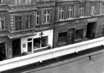 Blick auf die Galerie über die Mauer Hinweg. Am unteren Bildrand ist die weiße Mauerkrone zu sehen. Die Fassade rund um die Schaufenster der Galerie ist weiß gestrichen und sticht aus der ansonsten dunklen und beschädigten Fassade des Altbaus hervor.
