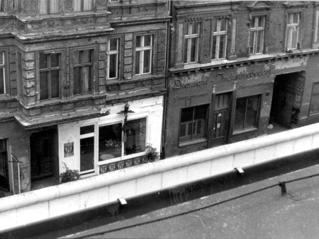 Blick auf die Galerie über die Mauer Hinweg. Am unteren Bildrand ist die weiße Mauerkrone zu sehen. Die Fassade rund um die Schaufenster der Galerie ist weiß gestrichen und sticht aus der ansonsten dunklen und beschädigten Fassade des Altbaus hervor.