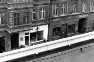 Blick auf die Galerie &uuml;ber die Mauer Hinweg. Am unteren Bildrand ist die wei&szlig;e Mauerkrone zu sehen. Die Fassade rund um die Schaufenster der Galerie ist wei&szlig; gestrichen und sticht aus der ansonsten dunklen und besch&auml;digten Fassade des Altbaus hervor.