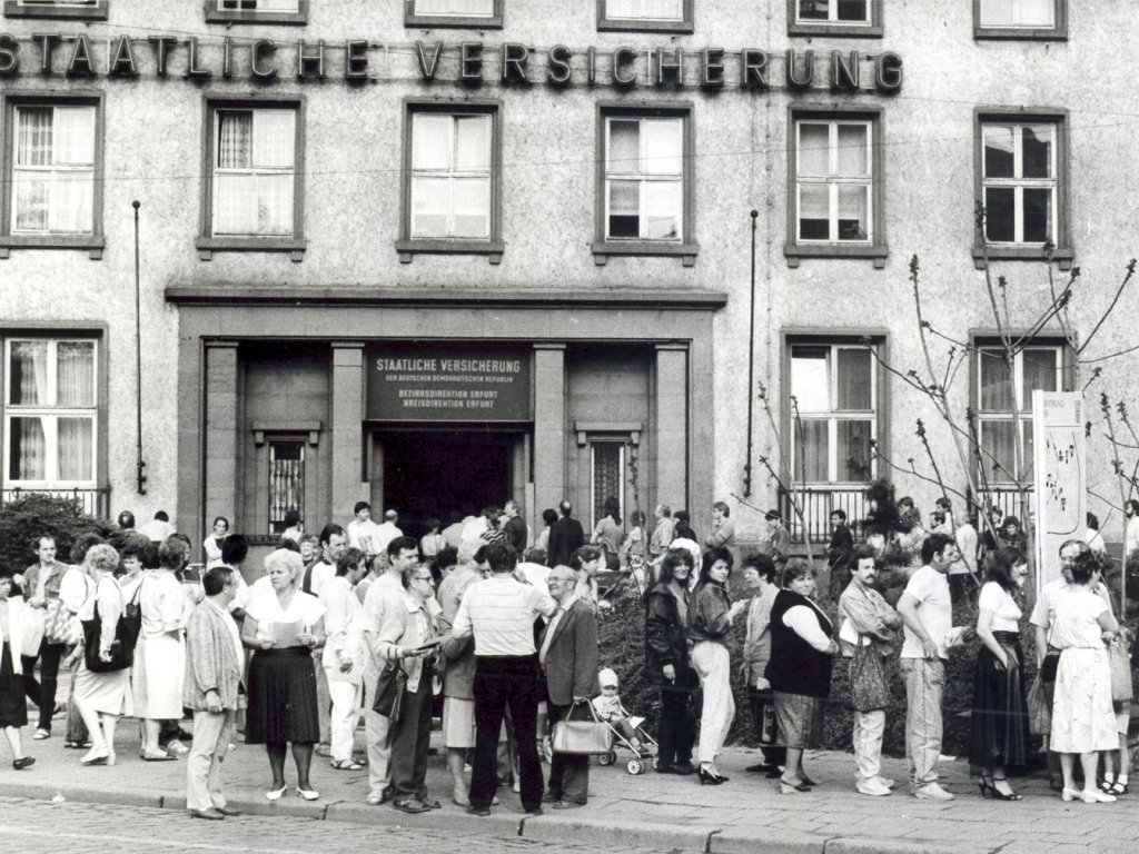Ansturm auf die staatliche Versicherung der DDR, nachdem bekannt wird, dass Versicherungen im Verhältnis 2:1 in D-Mark umgetauscht werden sollen, 8. Mai 1990. Zahlreiche Menschen stehen Schlange vor einem Gebäude mit der großen Fassadenbeschriftung Staatliche Versicherung. Einige stehen in Gruppen zusammen, halten Dokumente in den Händen und unterhalten sich. Über dem offenen Eingangstor steht Staatliche Versicherung der Deutschen Demokratischen Republik. Bezirksdirektion Erfurt. Kreisdirektion Erfurt.