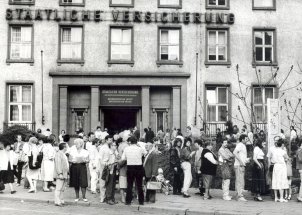 Zahlreiche Menschen stehen Schlange vor einem Gebäude mit der großen Fassadenbeschriftung Staatliche Versicherung. Einige stehen in Gruppen zusammen, halten Dokumente in den Händen und unterhalten sich. Über dem offenen Eingangstor steht Staatliche Versicherung der Deutschen Demokratischen Republik. Bezirksdirektion Erfurt. Kreisdirektion Erfurt.