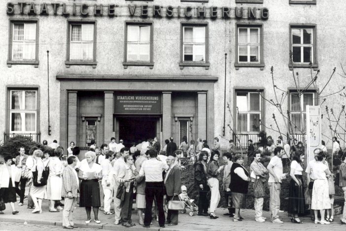 Zahlreiche Menschen stehen Schlange vor einem Gebäude mit der großen Fassadenbeschriftung Staatliche Versicherung. Einige stehen in Gruppen zusammen, halten Dokumente in den Händen und unterhalten sich. Über dem offenen Eingangstor steht Staatliche Versicherung der Deutschen Demokratischen Republik. Bezirksdirektion Erfurt. Kreisdirektion Erfurt.