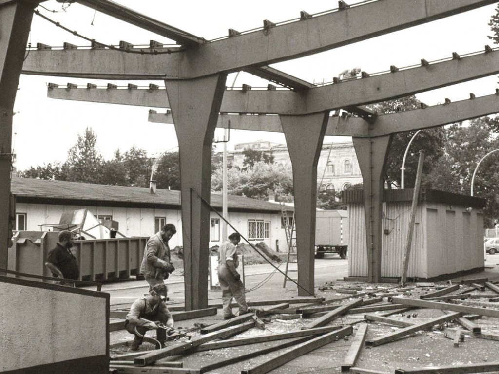 Das Dach des großen Unterstandes fehlt bereits, nur das Gerüst und zwei Baracken sind noch intakt. Auf dem Boden liegen verstreut Balken. Drei Männer stehen, einer ist in der Hocke und bearbeitet einen Balken mit einem Schweißgerät.