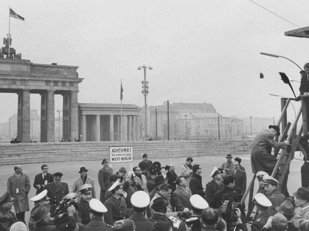 Im Bildhintergrund ist das Brandenburger Tor zu sehen, davor die Mauer. Uniformierte Männer und Journalisten drängen sich in der unteren Bildhälfte vor einem Schild mit der Aufschrift: Achtung, Sie verlassen jetzt West-Berlin. Rechts im Vordergrund steht ein Hochstand. Ein Mann klettert gerade die Leiter herauf, oben steht Robert Kennedy.