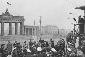 Im Bildhintergrund ist das Brandenburger Tor zu sehen, davor die Mauer. Uniformierte Männer und Journalisten drängen sich in der unteren Bildhälfte vor einem Schild mit der Aufschrift: Achtung, Sie verlassen jetzt West-Berlin. Rechts im Vordergrund steht ein Hochstand. Ein Mann klettert gerade die Leiter herauf, oben steht Robert Kennedy.