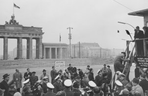 Im Bildhintergrund ist das Brandenburger Tor zu sehen, davor die Mauer. Uniformierte Männer und Journalisten drängen sich in der unteren Bildhälfte vor einem Schild mit der Aufschrift: Achtung, Sie verlassen jetzt West-Berlin. Rechts im Vordergrund steht ein Hochstand. Ein Mann klettert gerade die Leiter herauf, oben steht Robert Kennedy.