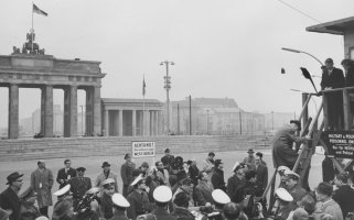 Im Bildhintergrund ist das Brandenburger Tor zu sehen, davor die Mauer. Uniformierte Männer und Journalisten drängen sich in der unteren Bildhälfte vor einem Schild mit der Aufschrift: Achtung, Sie verlassen jetzt West-Berlin. Rechts im Vordergrund steht ein Hochstand. Ein Mann klettert gerade die Leiter herauf, oben steht Robert Kennedy.