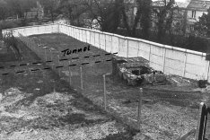 Vogelperspektive auf den Grenzstreifen von DDR-Seite aus. Links ein Teil eines Hauses. Davon ausgehend sind zwei gestrichelte Linien auf das Foto gemalt, die bis zur weißen Grenzmauer rechts gehen und beschriftet sind mit: Tunnel.