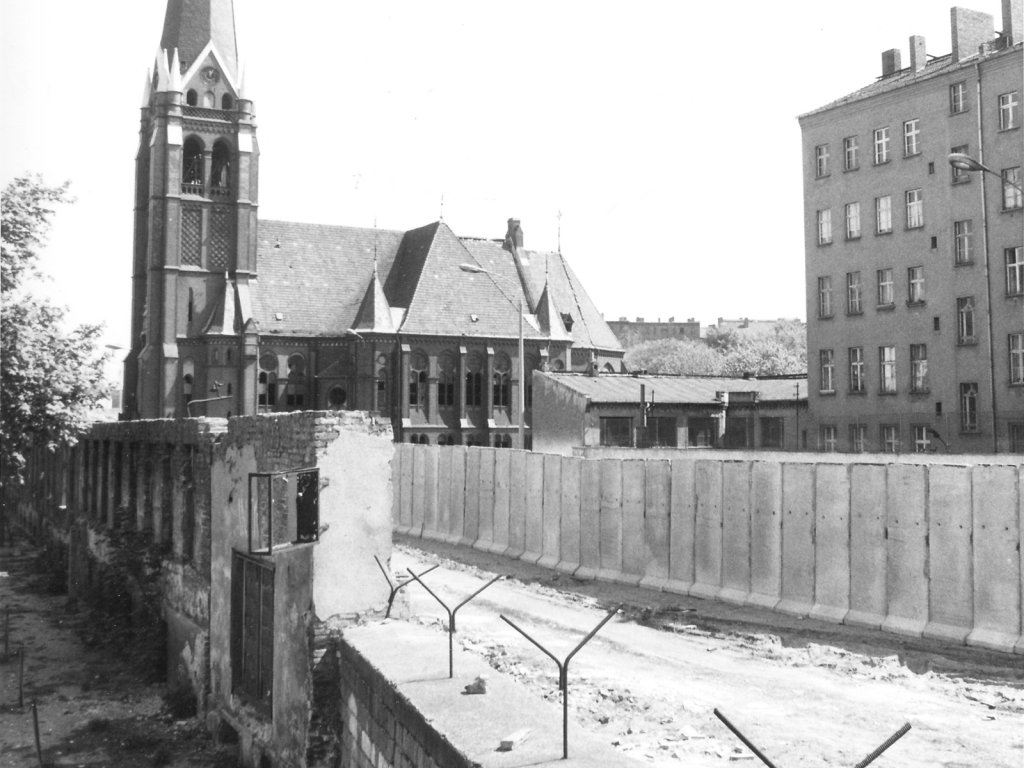 Mauer an der Bernauer Straße, im Hintergrund die Versöhnungskirche, 7. Juli 1980 Der Blick geht in den Grenzstreifen. Die Mauer besteht hier teilweise noch aus Fassadenresten abgerissener Grenzhäuser. Im Vordergrund sind Y-förmige Stacheldrahthalter auf der Mauerkrone.