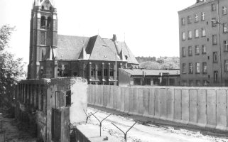Der Blick geht in den Grenzstreifen. Die Mauer besteht hier teilweise noch aus Fassadenresten abgerissener Grenzhäuser. Im Vordergrund sind Y-förmige Stacheldrahthalter auf der Mauerkrone.