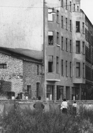 Ein Mann, eine Frau und ein Kind schauen über die Mauer, sie sind von hinten zu sehen. Auf der anderen Seite der Mauer steht eine Häuserzeile. Im zweiten Stock eines Wohnhauses stehen Menschen am Fenster.