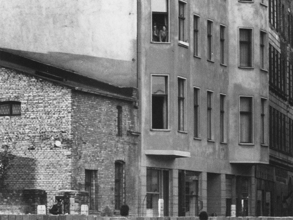 Ein Mann, eine Frau und ein Kind schauen über die Mauer, sie sind von hinten zu sehen. Auf der anderen Seite der Mauer steht eine Häuserzeile. Im zweiten Stock eines Wohnhauses stehen Menschen am Fenster.