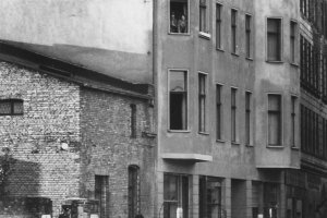 Ein Mann, eine Frau und ein Kind schauen über die Mauer, sie sind von hinten zu sehen. Auf der anderen Seite der Mauer steht eine Häuserzeile. Im zweiten Stock eines Wohnhauses stehen Menschen am Fenster.