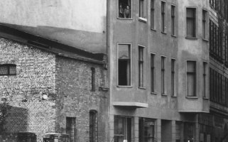 Ein Mann, eine Frau und ein Kind schauen über die Mauer, sie sind von hinten zu sehen. Auf der anderen Seite der Mauer steht eine Häuserzeile. Im zweiten Stock eines Wohnhauses stehen Menschen am Fenster.