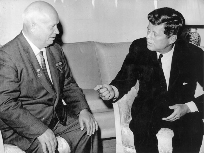 Links im Bild sitzt Nikita Chruschtschow, seine Hände liegen auf den Knien. Er schaut nach rechts zu John F. Kennedy, der gestikulierend auf ihn einredet.