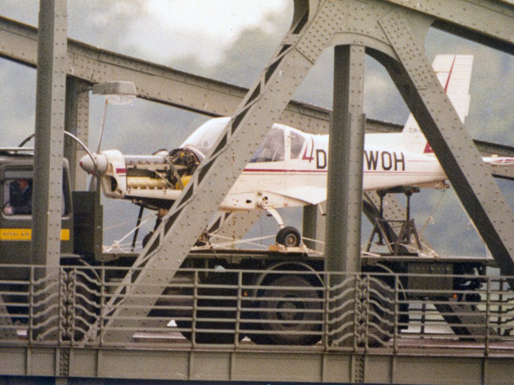 Rückgabe des zerlegten einmotorigen Flugzeugs durch britische Militärangehörige am 5. August 1987 an der Glienicker Brücke: Gelungene Flucht mit einem Motorflugzeug von Potsdam nach Berlin-Gatow, 15. Juli 1987 Ein weißes Kleinflugzeug steht auf der Lagefläche eines LKW. Im Vordergrund sind die Metallverstrebungen einer Brücke zu erkennen.