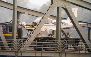 Ein weißes Kleinflugzeug steht auf der Lagefläche eines LKW. Im Vordergrund sind die Metallverstrebungen einer Brücke zu erkennen.
