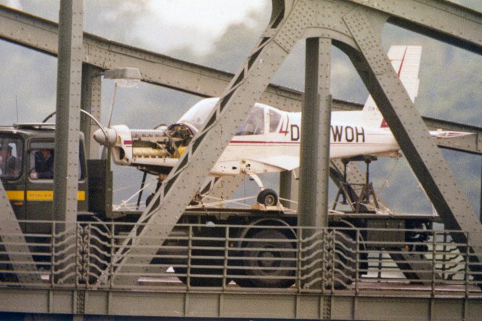Ein weißes Kleinflugzeug steht auf der Lagefläche eines LKW. Im Vordergrund sind die Metallverstrebungen einer Brücke zu erkennen.