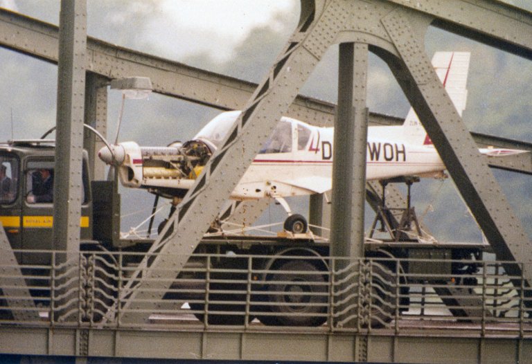 Ein weißes Kleinflugzeug steht auf der Lagefläche eines LKW. Im Vordergrund sind die Metallverstrebungen einer Brücke zu erkennen.