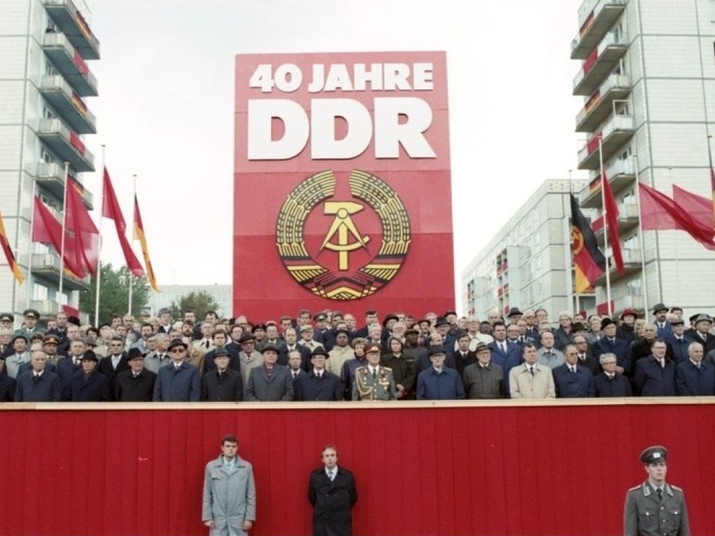 Auf einer rot verkleideten Tribüne stehen dutzende Menschen, die meisten von ihnen sind Männer in Anzügen, Hüten und Mänteln. Hinter ihnen steht ein riesiges rotes Plakat mit dem DDR-Staatsemblem und der weißen Aufschrift: 40 Jahre DDR. Links und rechts davon wehen DDR-Flaggen und rote Fahnen. Im Hintergrund sind die Wohnblöcke der Berliner Karl-Marx-Allee erkennbar.