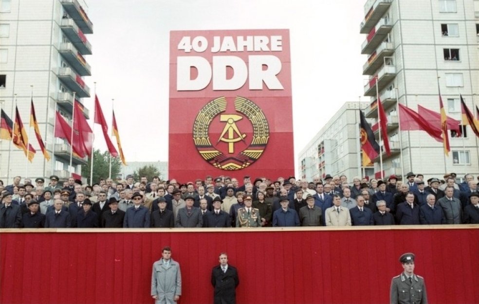 Auf einer rot verkleideten Tribüne stehen dutzende Menschen, die meisten von ihnen sind Männer in Anzügen, Hüten und Mänteln. Hinter ihnen steht ein riesiges rotes Plakat mit dem DDR-Staatsemblem und der weißen Aufschrift: 40 Jahre DDR. Links und rechts davon wehen DDR-Flaggen und rote Fahnen. Im Hintergrund sind die Wohnblöcke der Berliner Karl-Marx-Allee erkennbar.