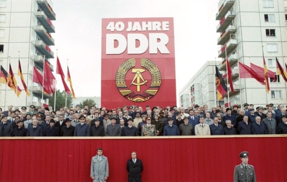 Auf einer rot verkleideten Tribüne stehen dutzende Menschen, die meisten von ihnen sind Männer in Anzügen, Hüten und Mänteln. Hinter ihnen steht ein riesiges rotes Plakat mit dem DDR-Staatsemblem und der weißen Aufschrift: 40 Jahre DDR. Links und rechts davon wehen DDR-Flaggen und rote Fahnen. Im Hintergrund sind die Wohnblöcke der Berliner Karl-Marx-Allee erkennbar.