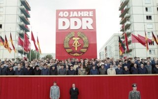 Auf einer rot verkleideten Tribüne stehen dutzende Menschen, die meisten von ihnen sind Männer in Anzügen, Hüten und Mänteln. Hinter ihnen steht ein riesiges rotes Plakat mit dem DDR-Staatsemblem und der weißen Aufschrift: 40 Jahre DDR. Links und rechts davon wehen DDR-Flaggen und rote Fahnen. Im Hintergrund sind die Wohnblöcke der Berliner Karl-Marx-Allee erkennbar.