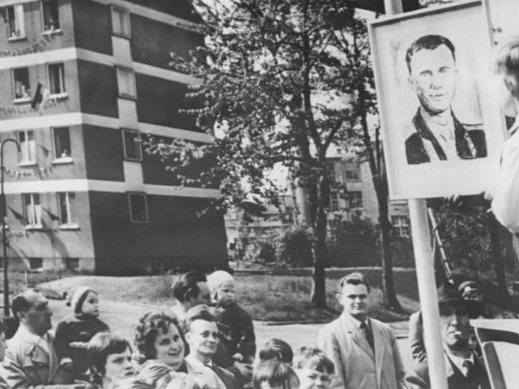 Im Vordergrund steht ein kleiner Junge auf einer Bühne neben einem gezeichneten Porträt von Juri Gagarin. Am unteren Bildrand sind einige Zuschauer zu sehen, im Bildhintergrund Bäume und ein Wohnhaus.