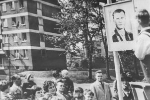 Im Vordergrund steht ein kleiner Junge auf einer Bühne neben einem gezeichneten Porträt von Juri Gagarin. Am unteren Bildrand sind einige Zuschauer zu sehen, im Bildhintergrund Bäume und ein Wohnhaus.