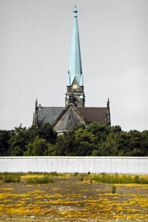 Auf dem Grenzstreifen im Vordergrund blühen gelbe Lupinen. Dahinter ist ein Stück Berliner Mauer zu sehen und im Hintergrund steht die Sanktsebastiankirche.