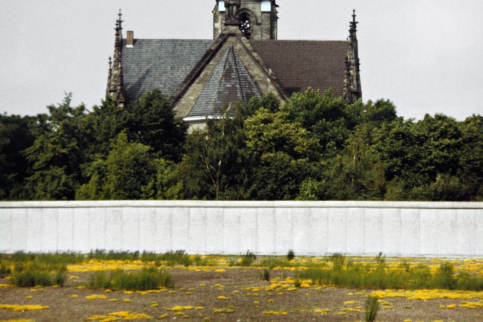 Auf dem Grenzstreifen im Vordergrund blühen gelbe Lupinen. Dahinter ist ein Stück Berliner Mauer zu sehen und im Hintergrund steht die Sanktsebastiankirche.