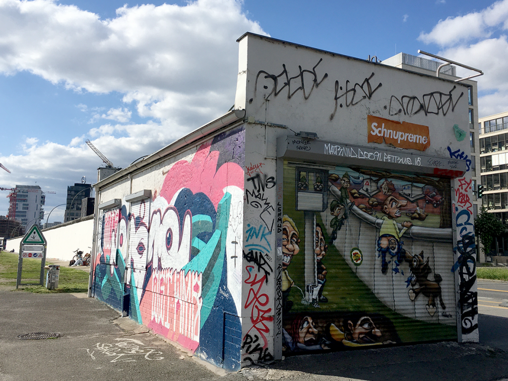 Zentral im Bild steht ein kleines Gebäude, das mit Graffiti buntbemalt ist. Der Eingang auf der Vorderseite und zwei Fenster auf der rechten Seite sind mit Rollläden verschlossen. Im linken Bildhintergrund ist die weißbemalte, der Spree zugewandte Rückseite der East Side Gallery zu sehen, am rechten Bildrand erkennt man die Mühlenstraße.