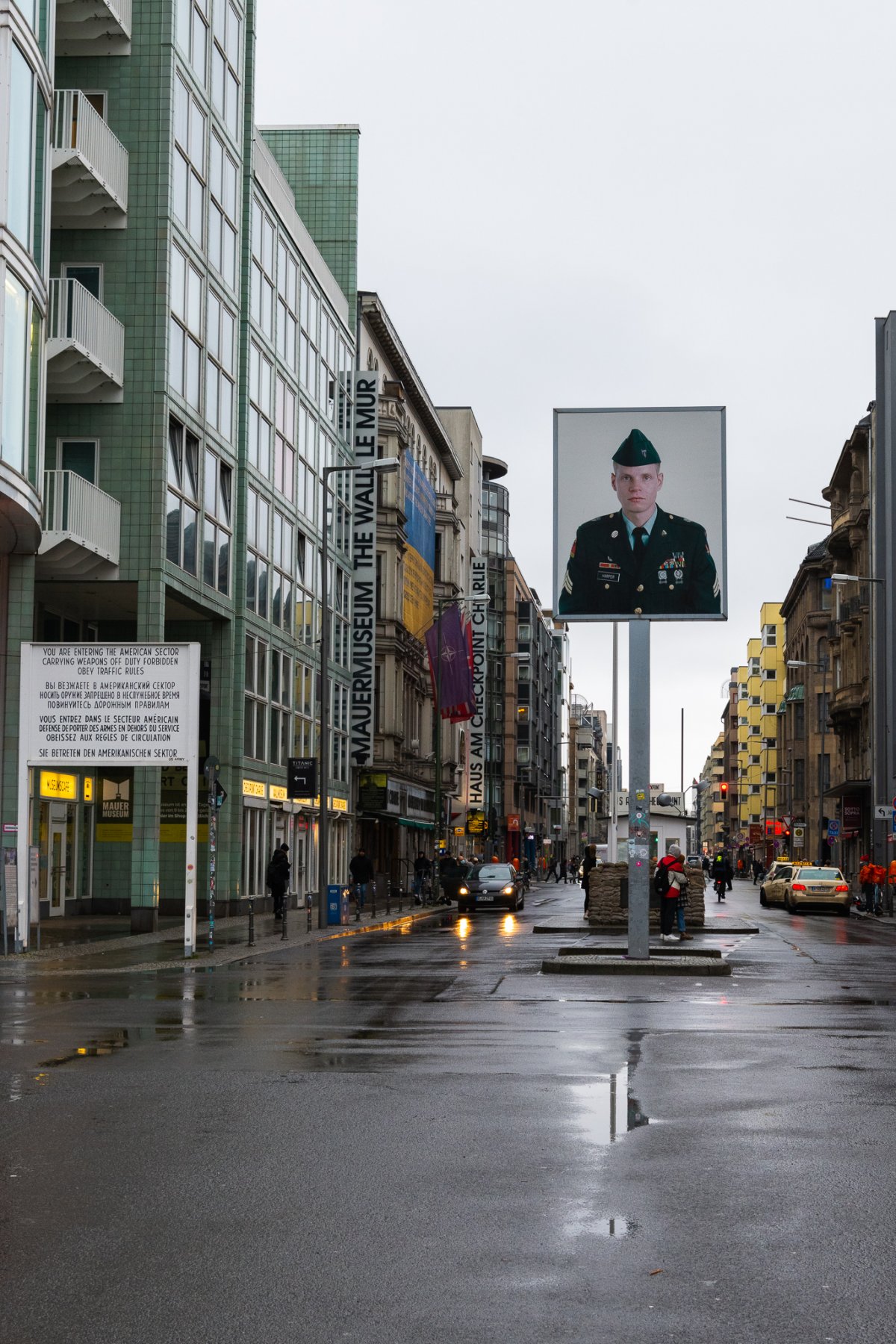 Checkpoint Charlie an einem regnerischen Tag, links Sektorenschild mit Aufschrift: You are entering the american sector. Carrying weapons off durty forbidden. Obey traffic rules. Mittig Mauermuseum, rechts Kontrollbaracke mit Sandsäcken, davor eine Verkehrsinsel mit Leuchtkasten in einigen Metern Höhe, zeigt überlebensgroßes Foto eines amerikanischen Soldaten. Checkpoint Charlie an einem regnerischen Tag, links Sektorenschild mit Aufschrift: You are entering the american sector. Carrying weapons off durty forbidden. Obey traffic rules. Mittig Mauermuseum, rechts Kontrollbaracke mit Sandsäcken, davor eine Verkehrsinsel mit Leuchtkasten in einigen Metern Höhe, zeigt überlebensgroßes Foto eines amerikanischen Soldaten.