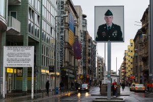 Checkpoint Charlie an einem regnerischen Tag, links Sektorenschild mit Aufschrift: You are entering the american sector. Carrying weapons off durty forbidden. Obey traffic rules. Mittig Mauermuseum, rechts Kontrollbaracke mit Sandsäcken, davor eine Verkehrsinsel mit Leuchtkasten in einigen Metern Höhe, zeigt überlebensgroßes Foto eines amerikanischen Soldaten.