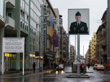 Checkpoint Charlie an einem regnerischen Tag, links Sektorenschild mit Aufschrift: You are entering the american sector. Carrying weapons off durty forbidden. Obey traffic rules. Mittig Mauermuseum, rechts Kontrollbaracke mit Sandsäcken, davor eine Verkehrsinsel mit Leuchtkasten in einigen Metern Höhe, zeigt überlebensgroßes Foto eines amerikanischen Soldaten.