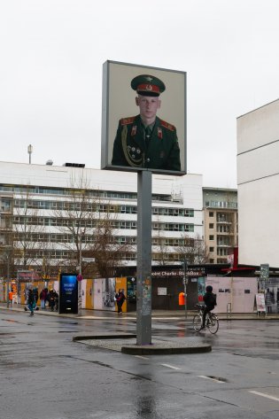 Checkpoint Charlie on a rainy day. In the center of the photo is the lightbox rising above the street. It shows a larger than scale photo of a russian soldier. In the background is an intersection and the Checkpoint Charlie BlackBox.